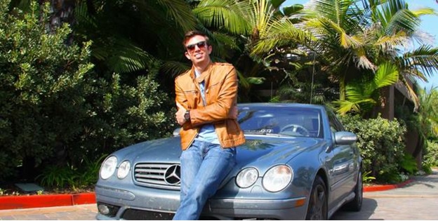 Man in a brown jacket leaning on the hood of a blue Mercedes car.