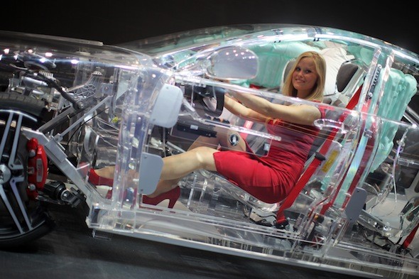 Woman in a red dress behind the wheel of a see through car.