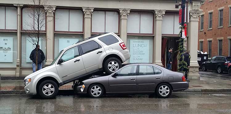 White SUV parked on top of a small grey 4-door car.