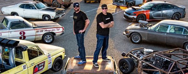 Two guys standing back to back on the hood of a car surrounded by souped up muscle cars.