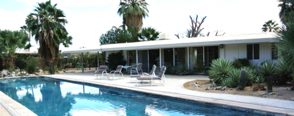 A one story house with a fancy pool behind it; pool chairs layed out by the pool.